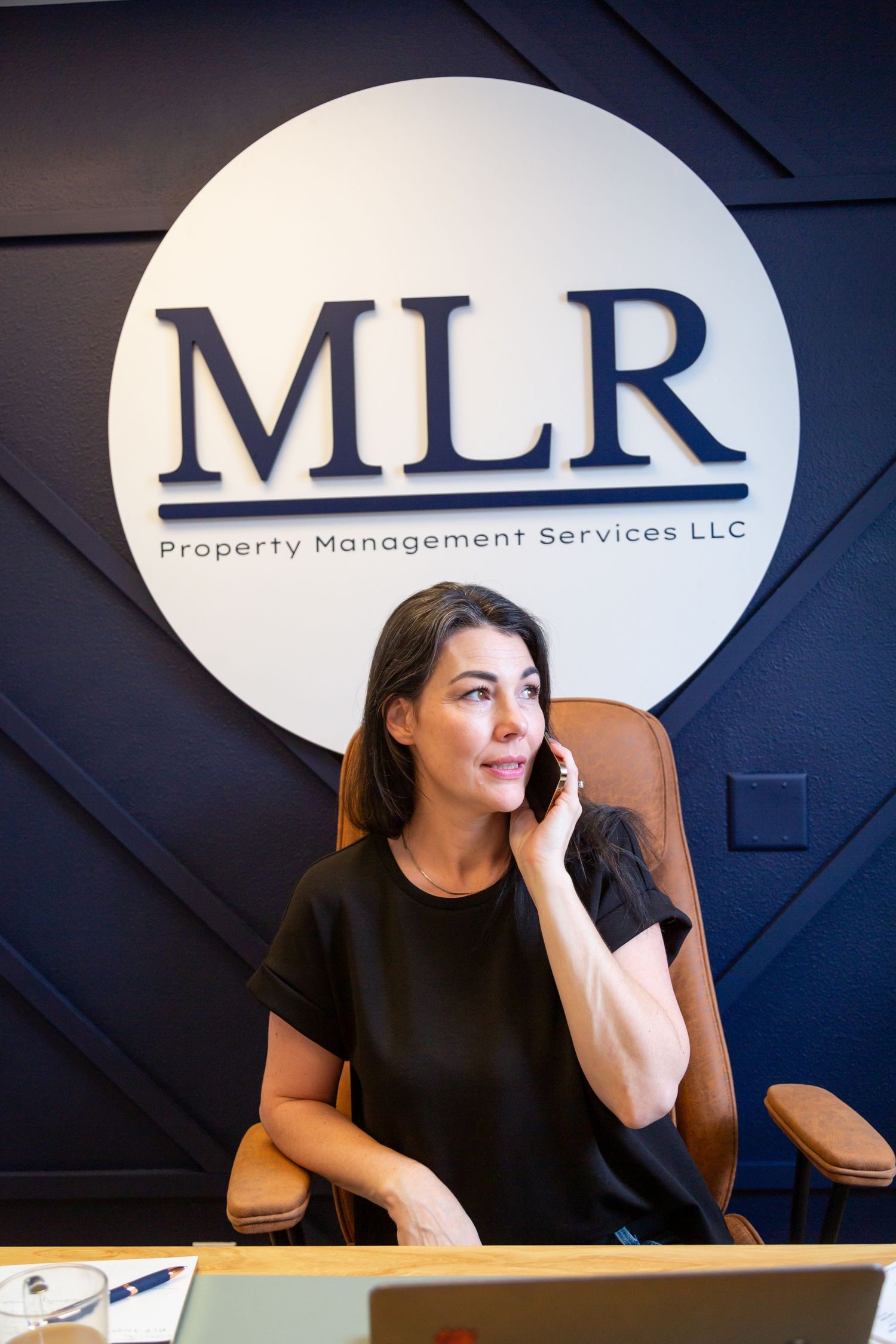 A professional woman in a black top talks on the phone at a desk in front of an MLR Property Management Services LLC sign.