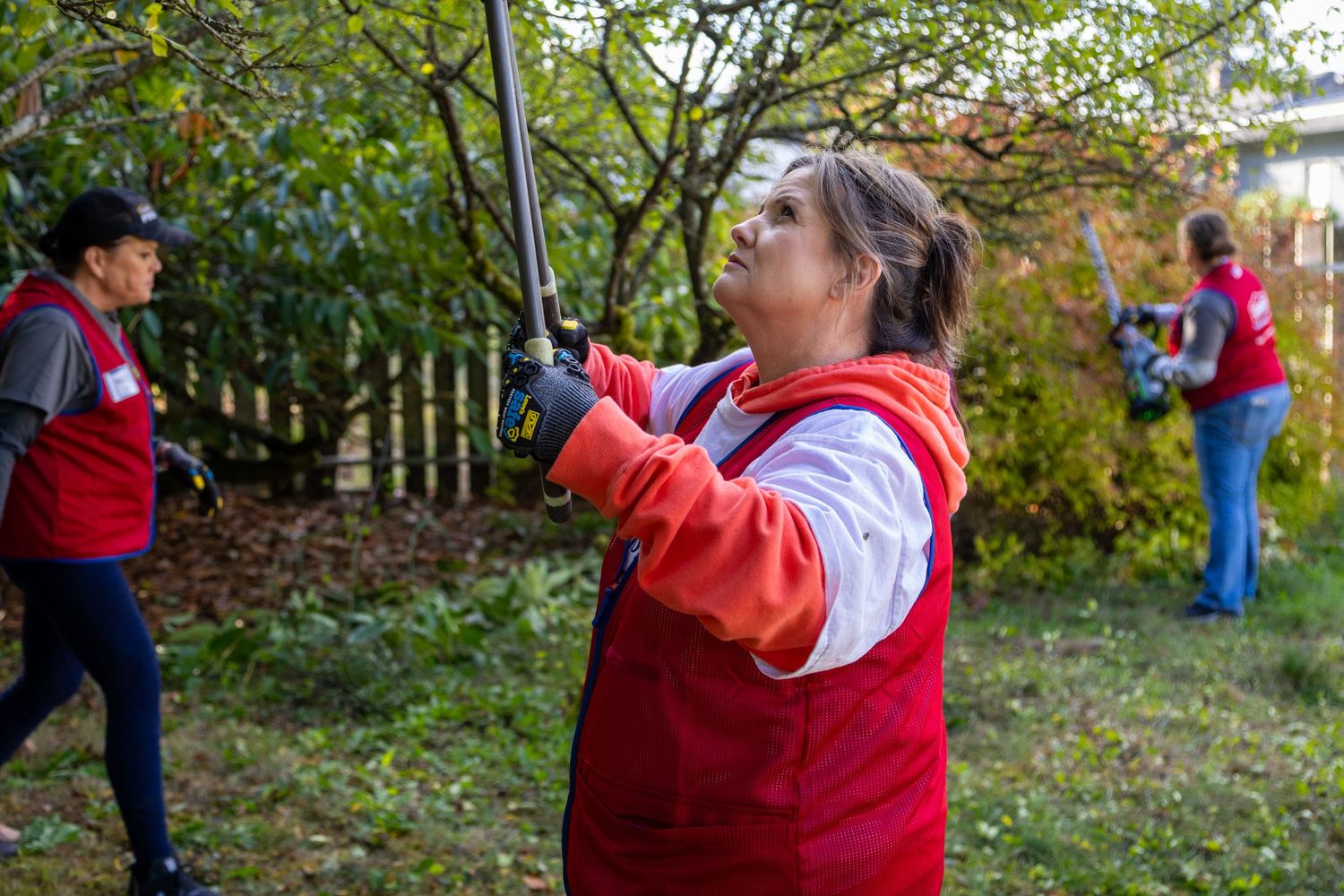 Volunteers in red vests trimming hedges and doing yard work outdoors during a community service event.