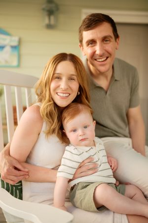 A smiling couple poses with their red-haired baby boy on a cozy porch setting.