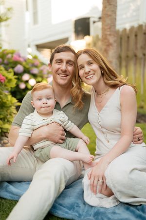 Happy young family with baby sitting on blanket in garden with pink flowers.