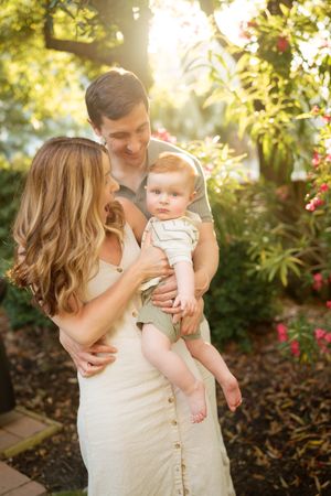 Young couple holding baby in sunlit garden with lush greenery and pink flowers.