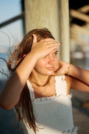 Young woman shielding eyes from golden sunset light near wooden pier, wearing white smocked top and gold jewelry.