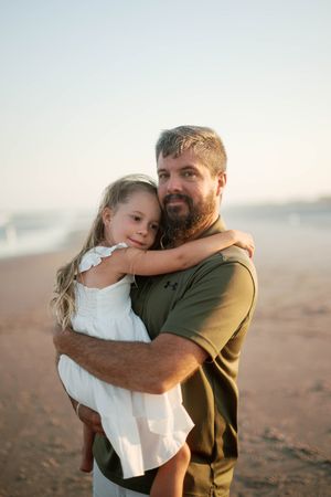 A bearded father holds his young daughter in a white dress on a sandy beach at golden hour.