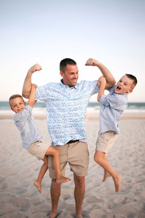 Father flexing muscles on beach while holding two laughing young boys.
