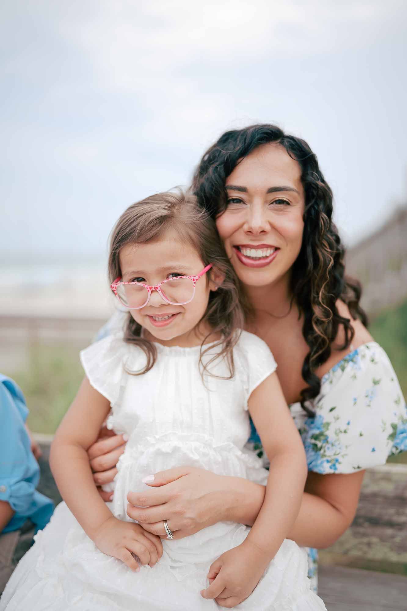 A smiling woman with curly hair hugs a young girl in pink glasses and white dress at a beach.