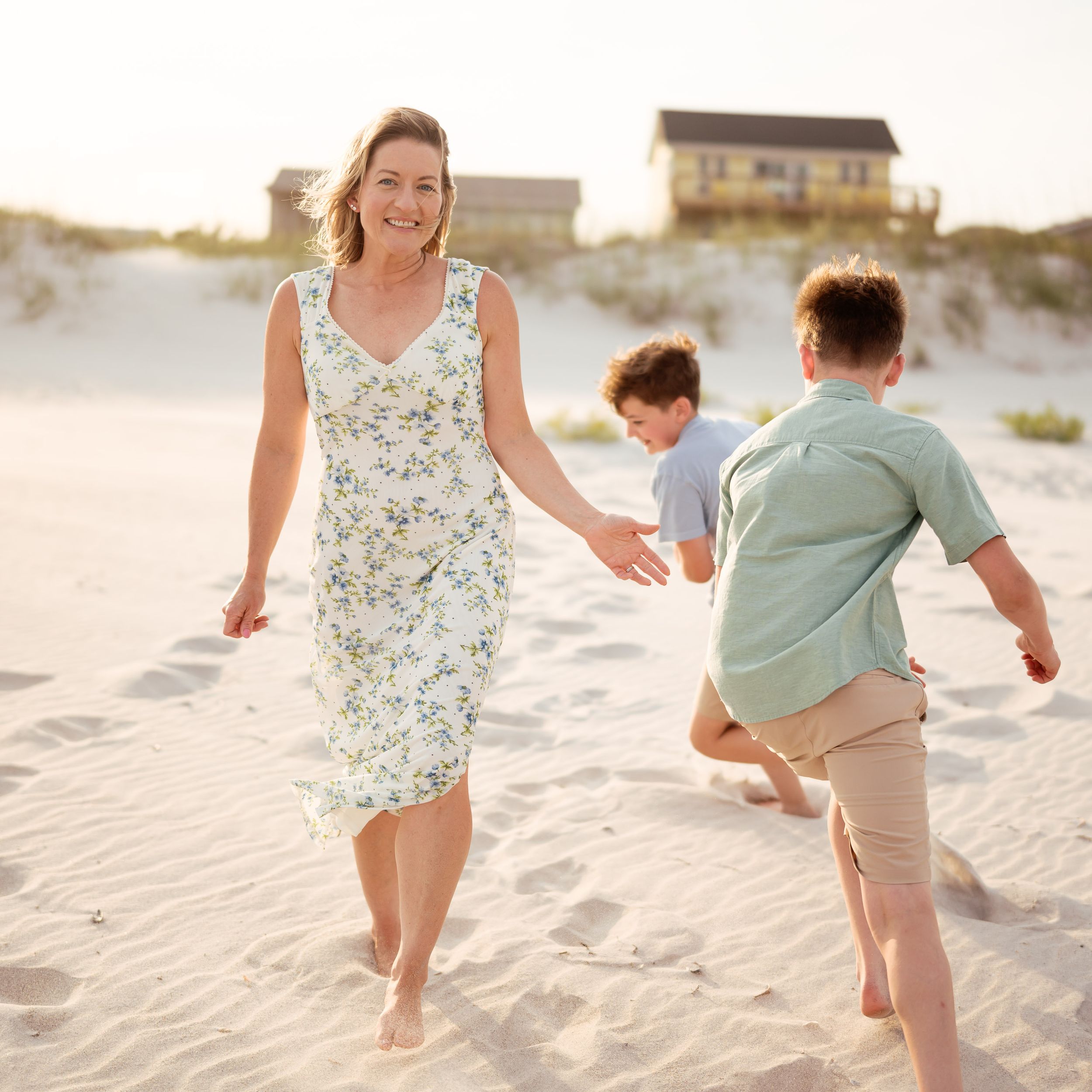 A woman in a floral dress walks on a sunny beach with two boys playing in the sand near coastal houses.