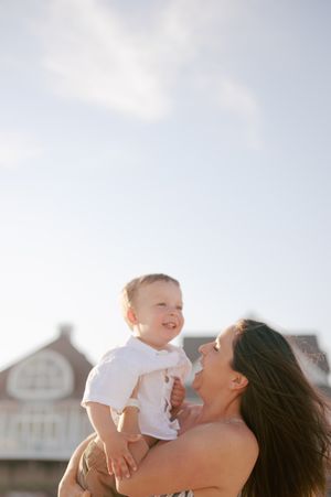 Mother holds smiling baby boy up against a bright blue sky near beachside buildings.