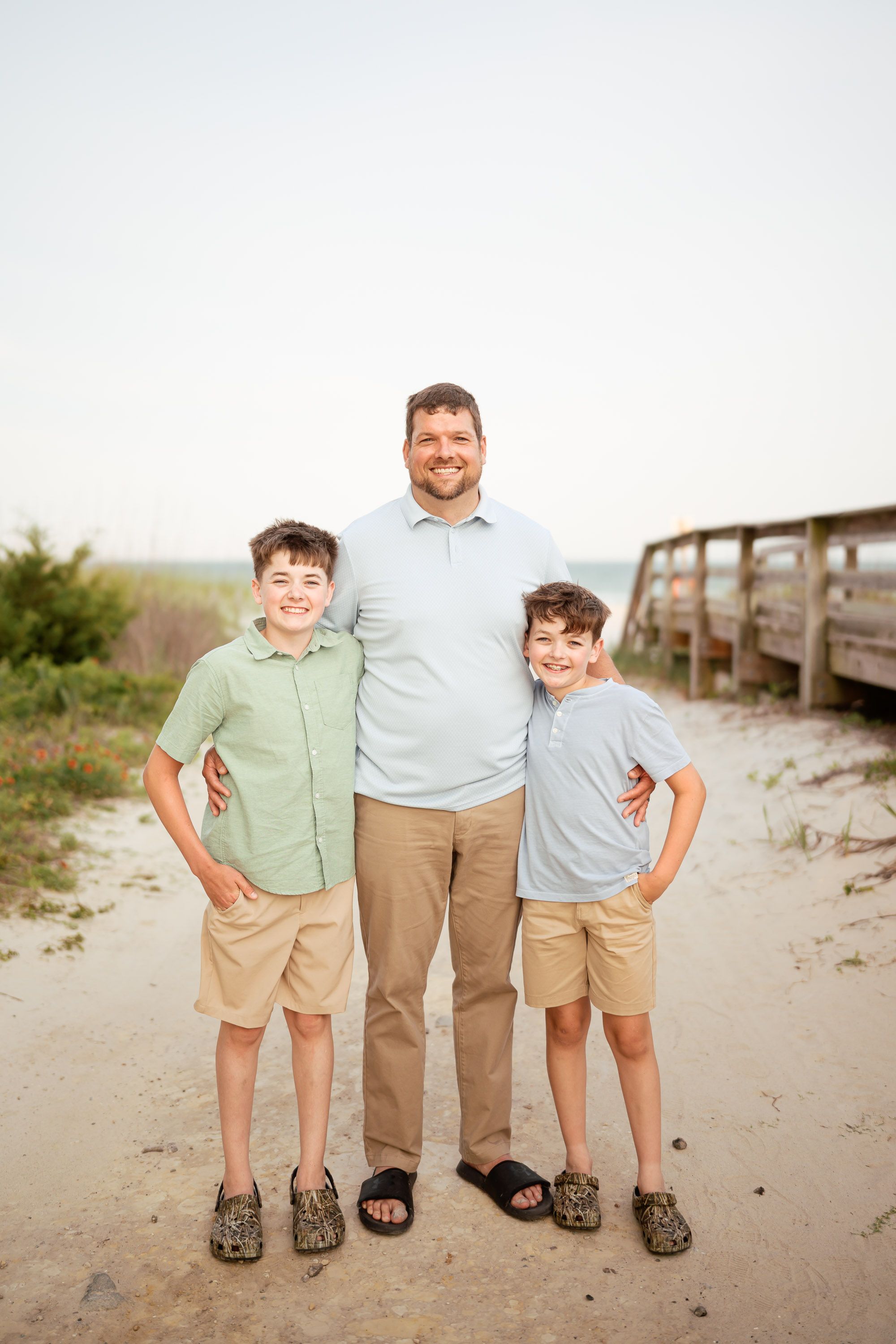 A smiling father poses with his two young sons on a sandy beach path near a wooden boardwalk.