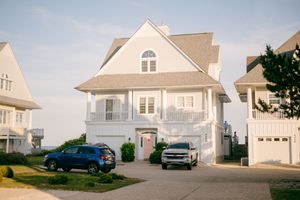 A large white two-story beach house with balcony and two vehicles parked in the driveway.