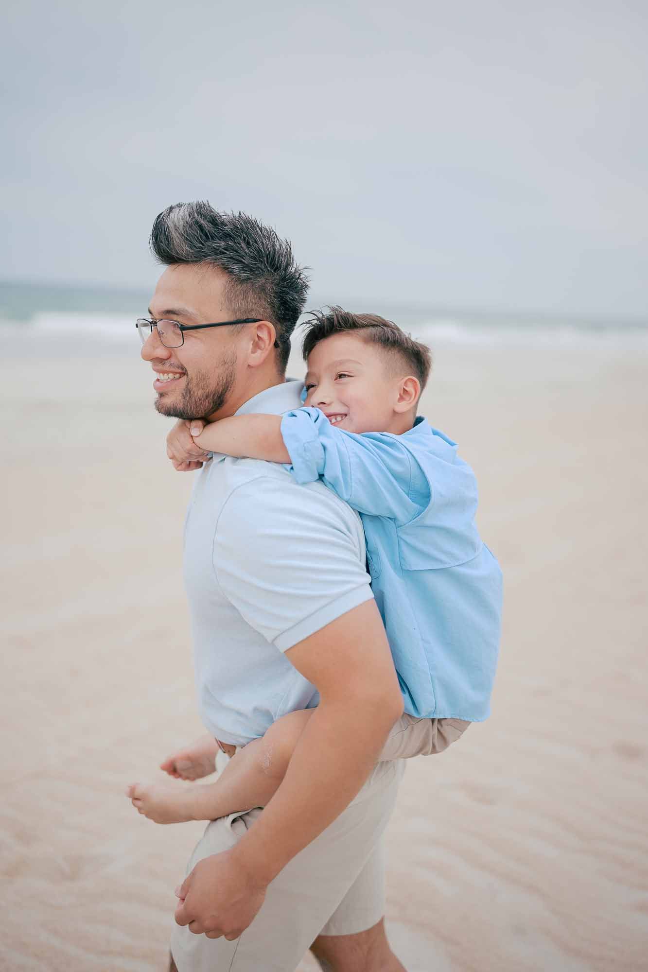 A smiling man gives a laughing young boy a piggyback ride on a sandy beach by the ocean.