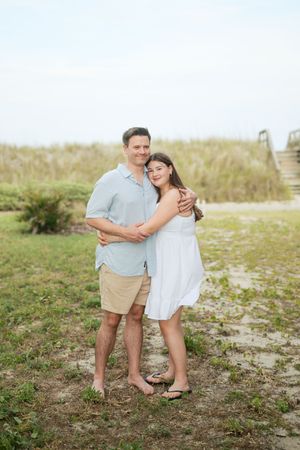 Father and teenage daughter embrace while standing barefoot near beach dunes on overcast day.