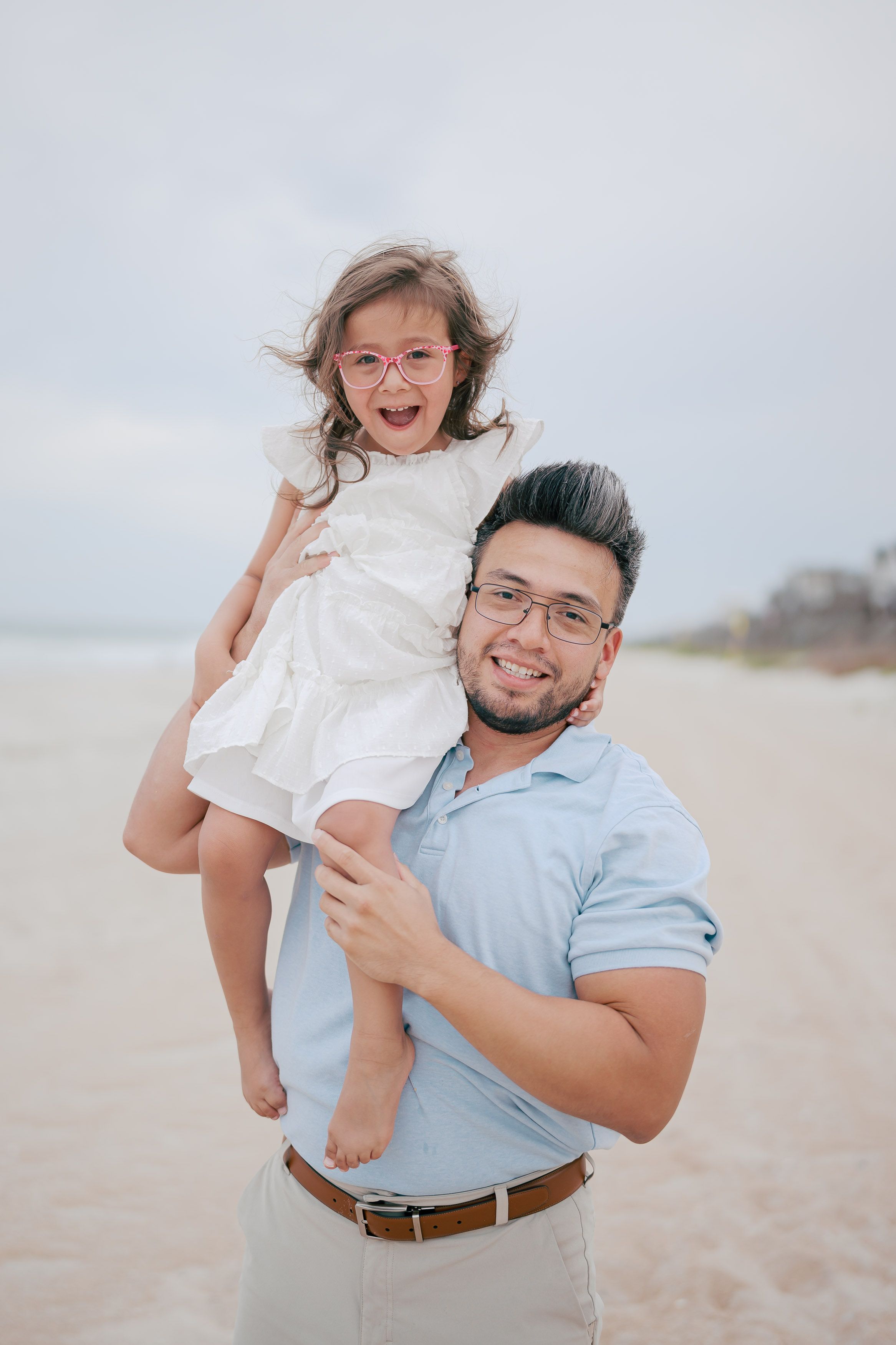 A joyful father holds his laughing little girl in a white dress and pink glasses on a sandy beach.