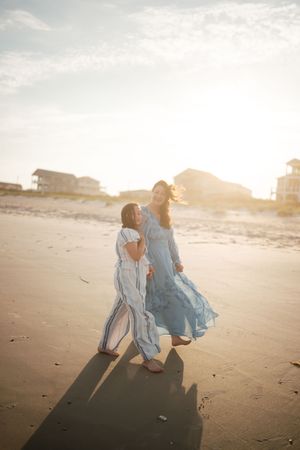 Two women in flowing blue dresses walking on sunny beach at golden hour with coastal homes in background.