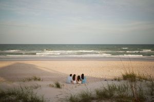 Four people sit together on a sandy beach facing calm ocean waves at dusk with sea oats in the foreground.