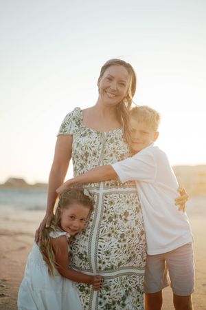 Pregnant mother smiling on beach at sunset with son and daughter hugging her.