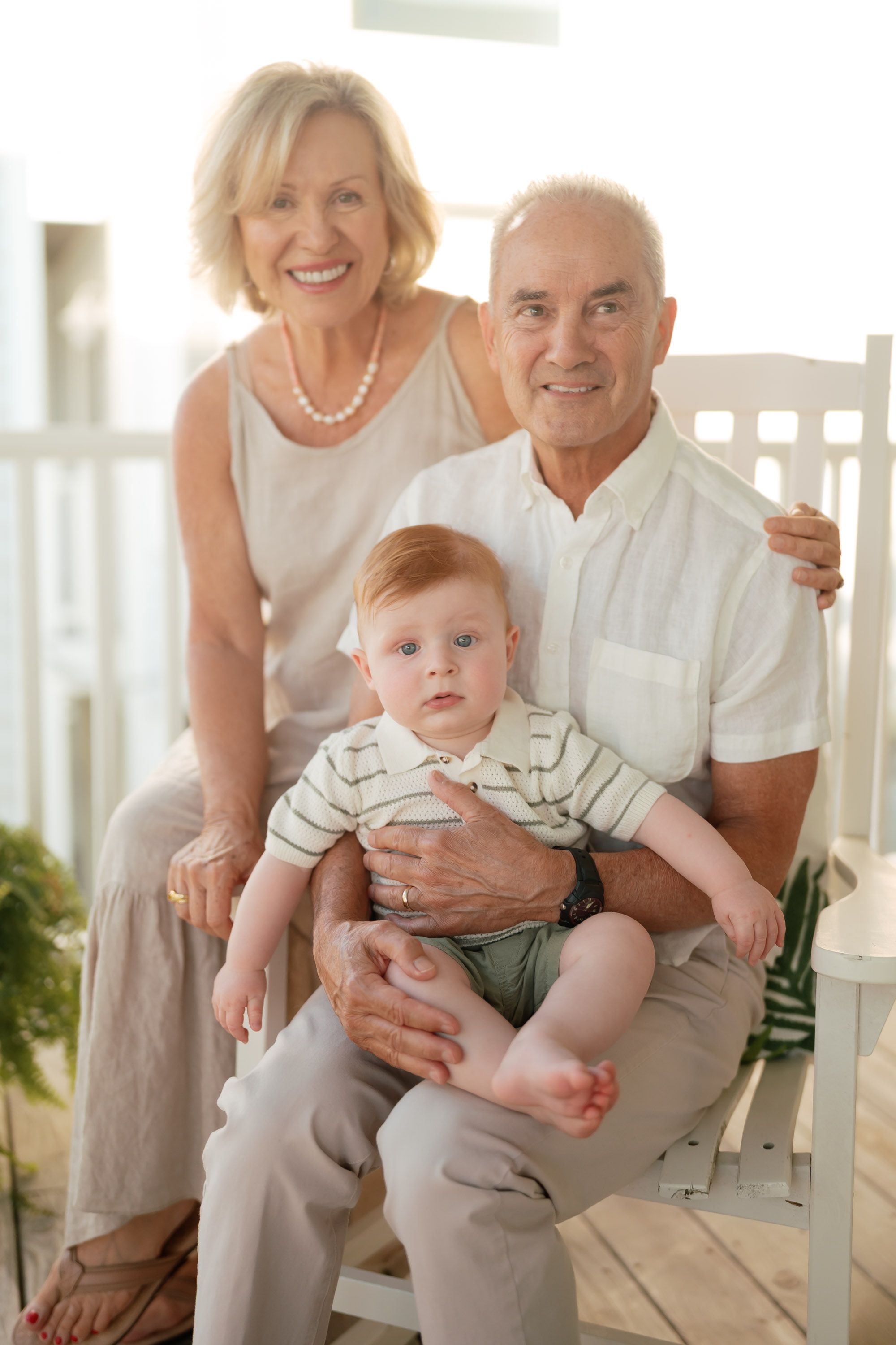 Grandparents smiling on white bench while holding a red-haired baby on porch.