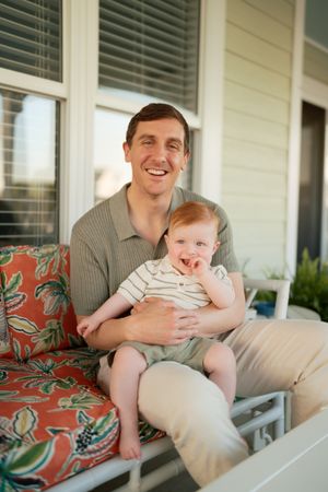 Smiling father sitting on porch swing holding happy red-haired baby on floral cushioned outdoor sofa.