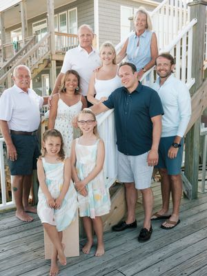 A cheerful family group poses on a beach house deck, dressed in summer whites and pastels, with ocean views behind them.