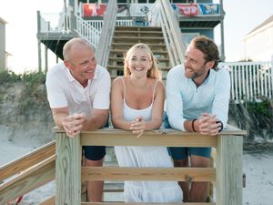 Three smiling adults pose at a wooden beach table with ocean stairs and a coastal house in the background.