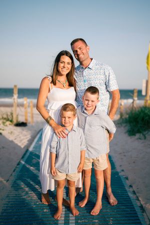 Family of four poses on a beach boardwalk at sunset, parents and two young boys in coordinating outfits.