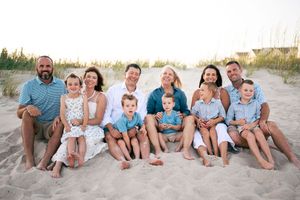 Large family group portrait on sandy beach, smiling adults and children sitting together in coordinated blue and white outfits.
