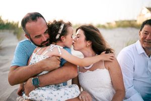 Parents lovingly kissing their young daughter on a beach while another family member looks on nearby.