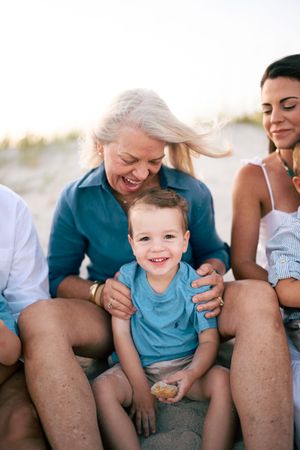 Grandmother laughs with toddler grandson on sandy beach while mother smiles nearby at golden hour.