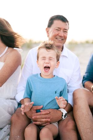 Young boy laughs joyfully while sitting on grandfather's lap during a sunny family beach session.