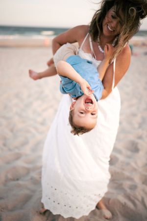 A laughing mother playfully holds her giggling toddler upside down on a sunny sandy beach.