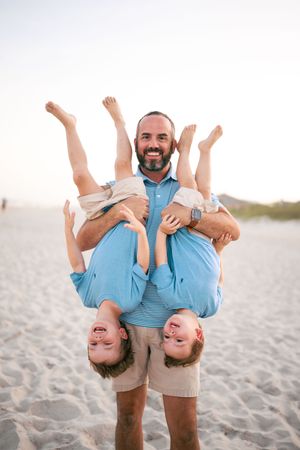 Smiling bearded dad holds two giggling boys upside down on a sunny sandy beach.