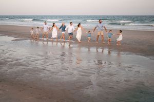 Large family holding hands walking on a sandy beach at sunset wearing coordinated white and beige outfits.