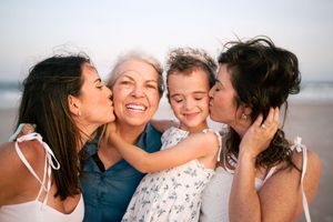 Four generations of women sharing joyful kisses and hugs on a sunny beach.