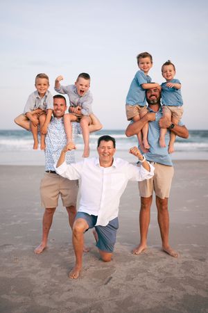 Three men pose playfully on beach carrying four young boys on their shoulders and back.