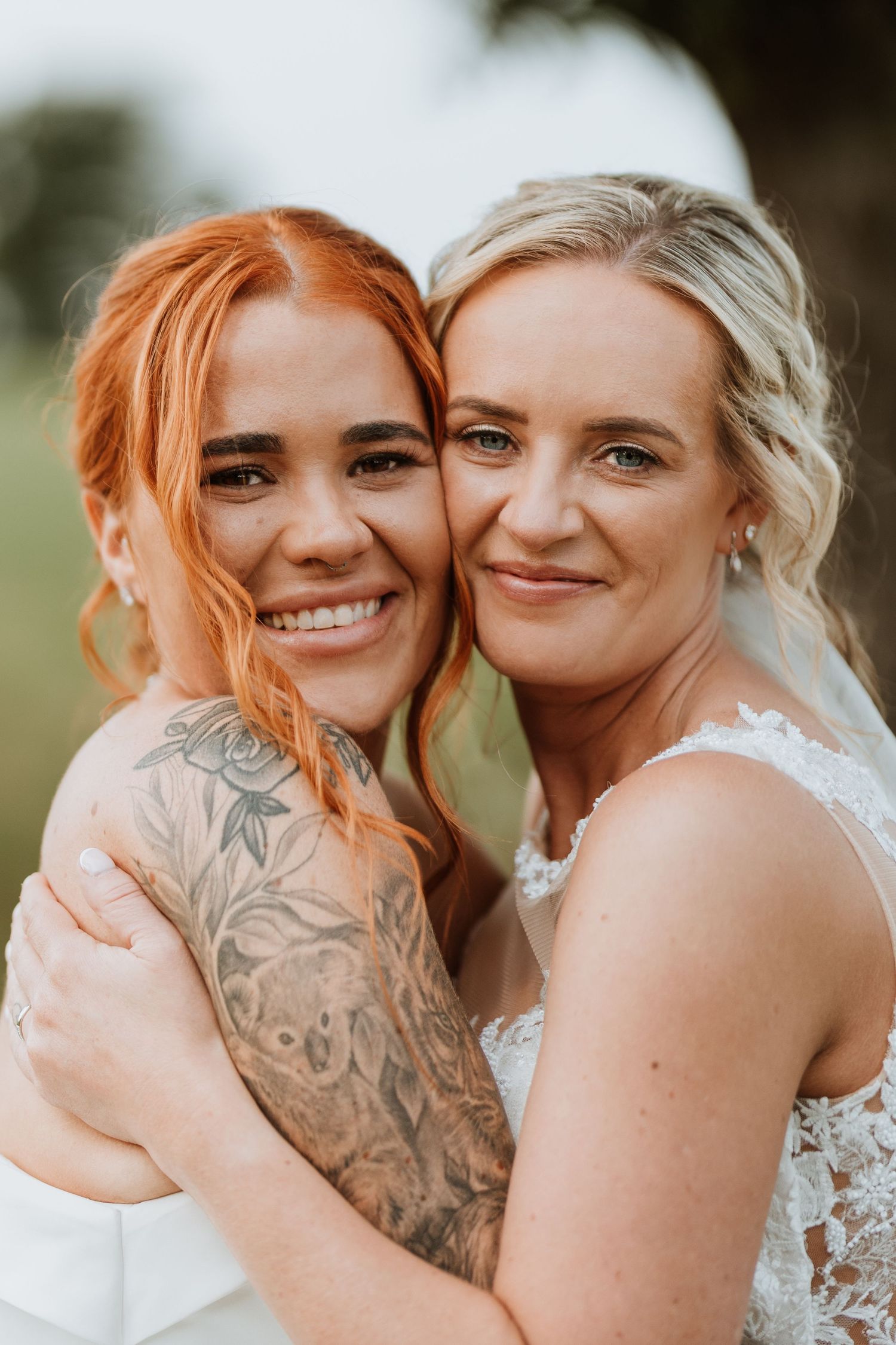 Two brides in white wedding dresses embrace outdoors, one with red hair and floral tattoos, the other with blonde curls.