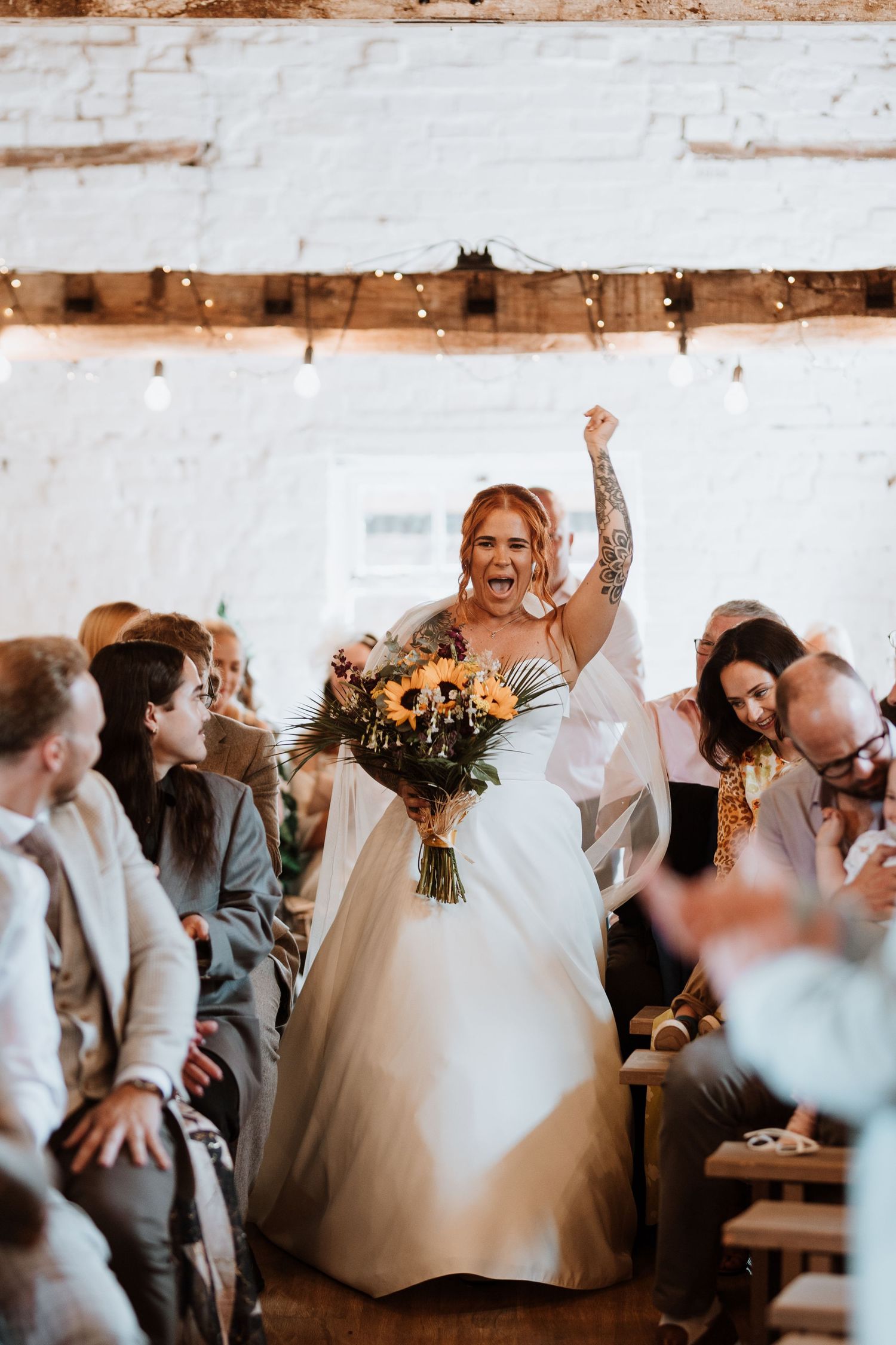 Joyful bride walks down the aisle raising her fist, holding sunflower bouquet, in a rustic white brick venue.