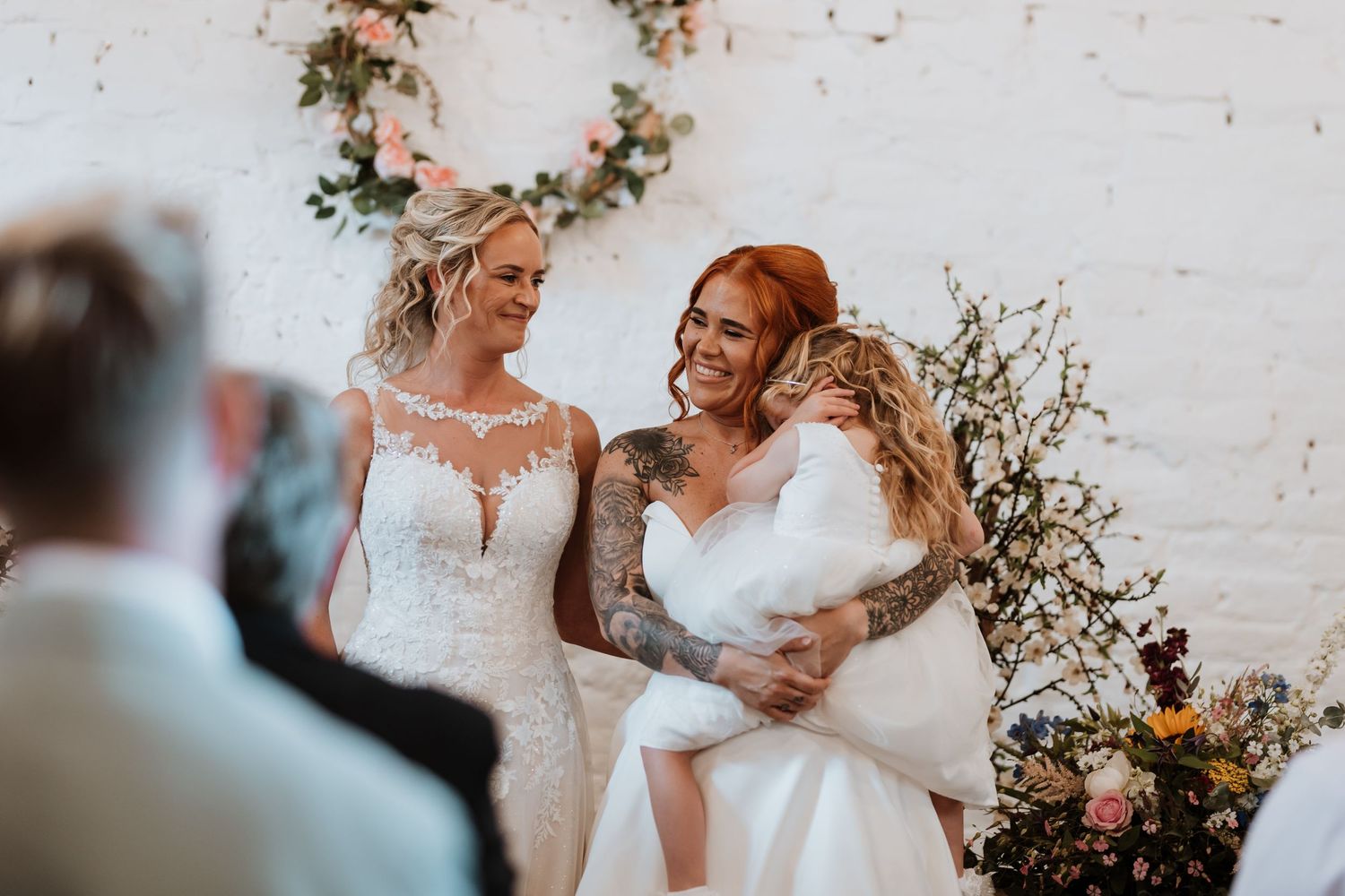 Two brides in white wedding dresses smiling and embracing during their ceremony in front of a floral arch.