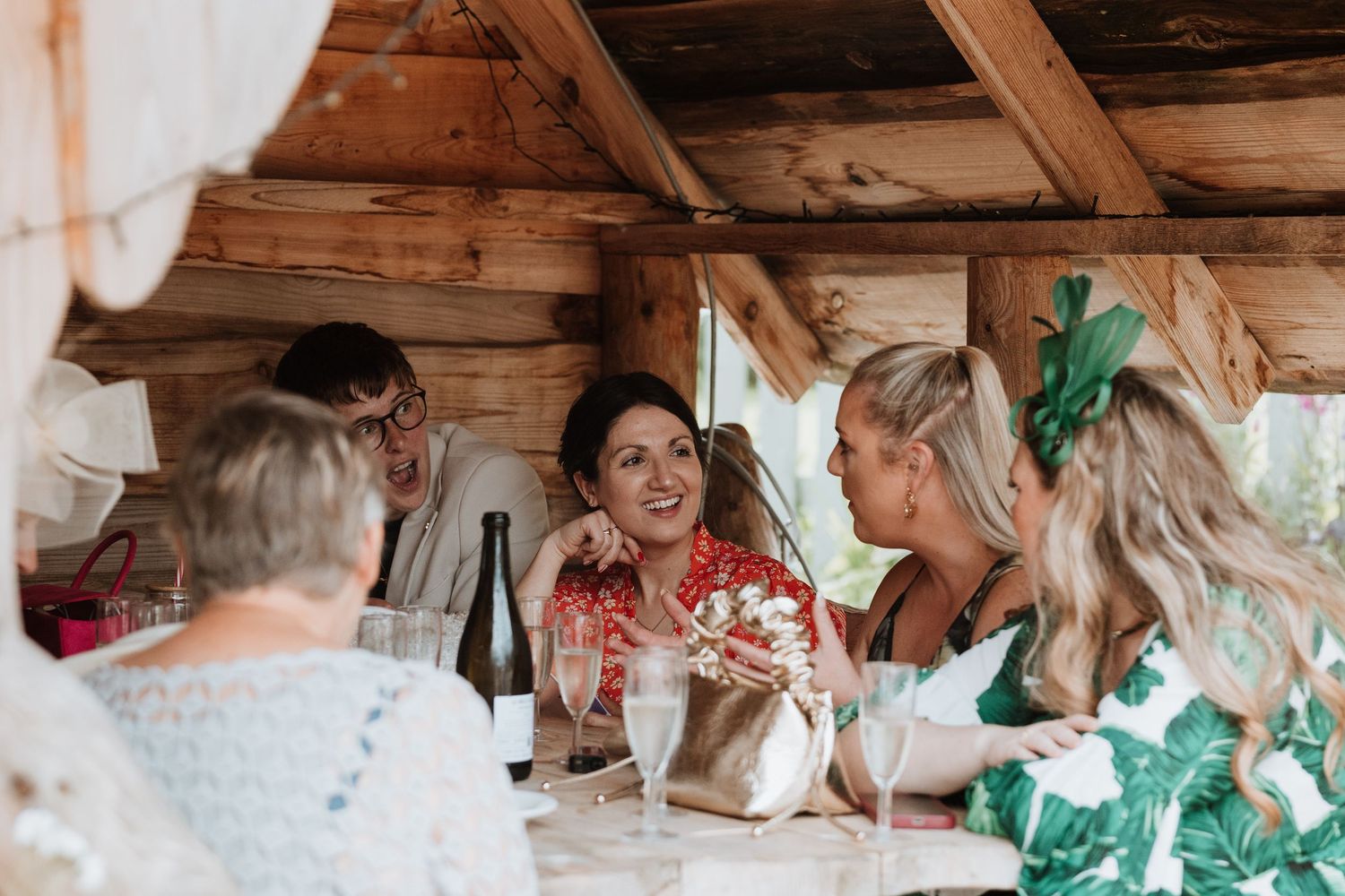 Group of women laughing and chatting at a rustic wooden cabin table during a wedding celebration.