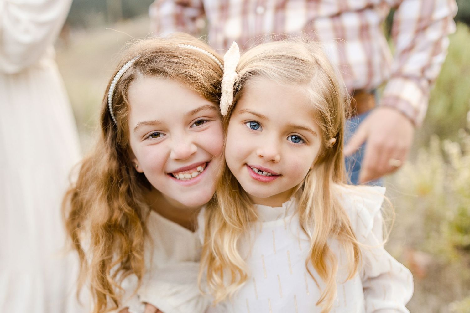 Two young sisters smiling together outdoors, wearing white dresses with hair accessories in a candid family portrait.
