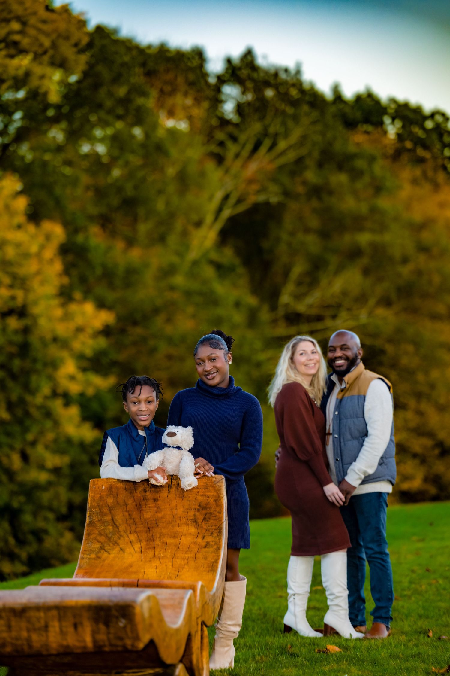 Family of four posing outdoors in autumn park with wooden sled and teddy bear, surrounded by colorful fall foliage.