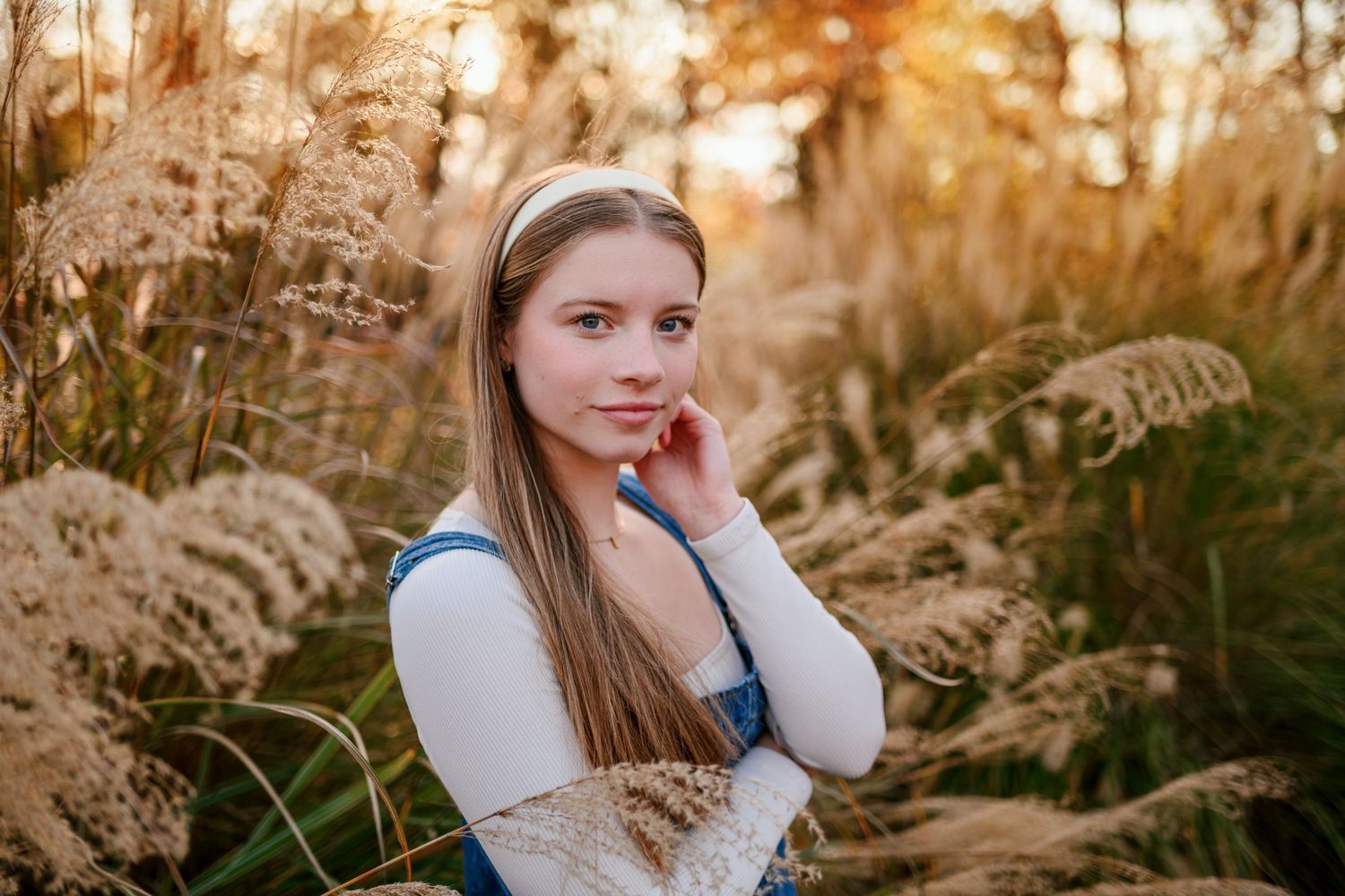 A teenage girl with long hair and a white headband poses among golden ornamental grasses in warm autumn light.