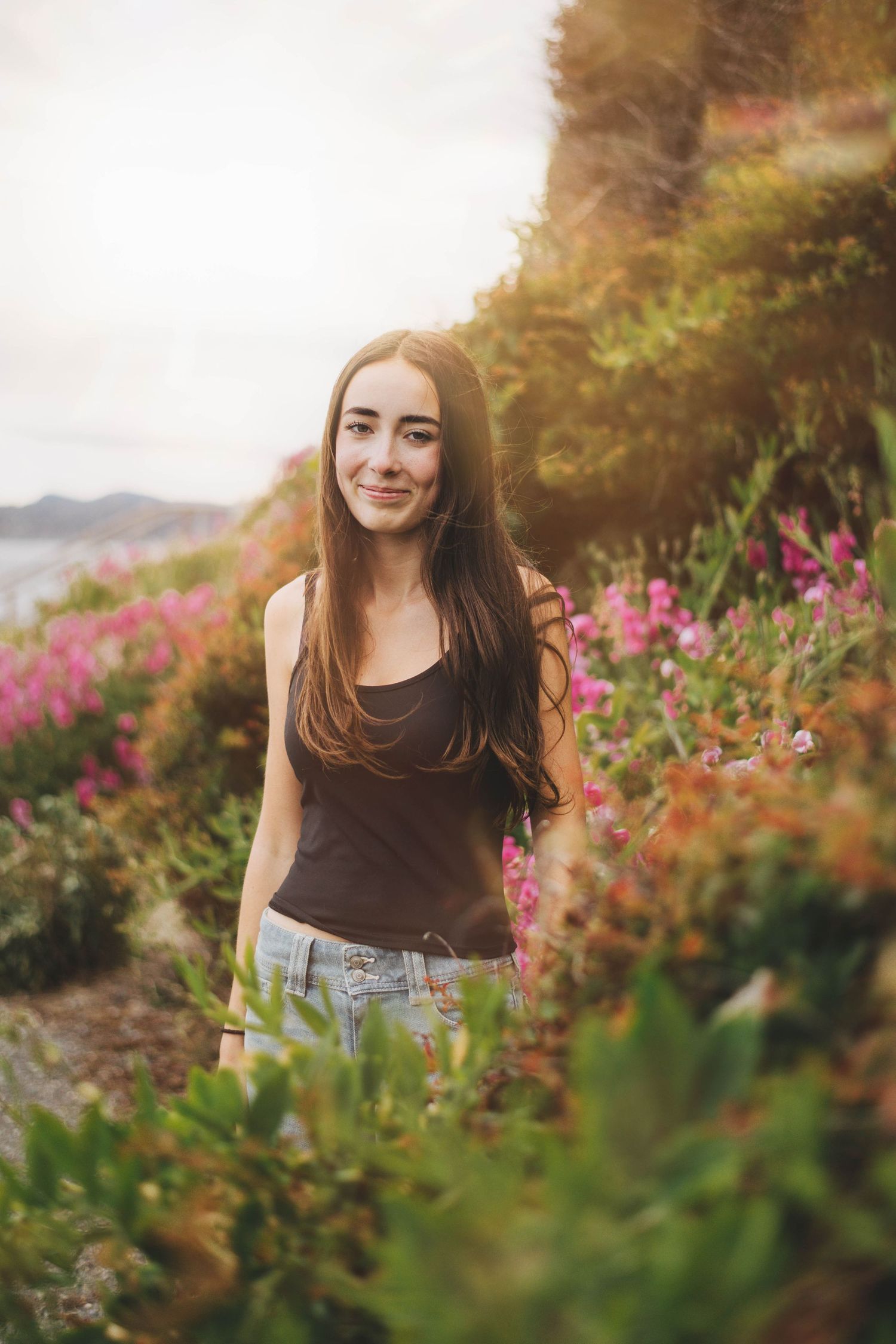 Smiling pregnant woman in pink off-shoulder dress with braided hair, standing outdoors in golden sunlight.