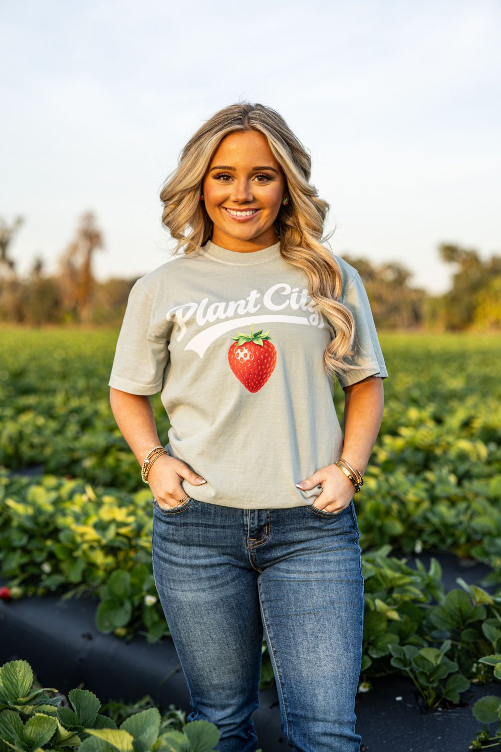Woman wearing a sage green Plant City strawberry graphic tee with jeans, standing in a strawberry field.