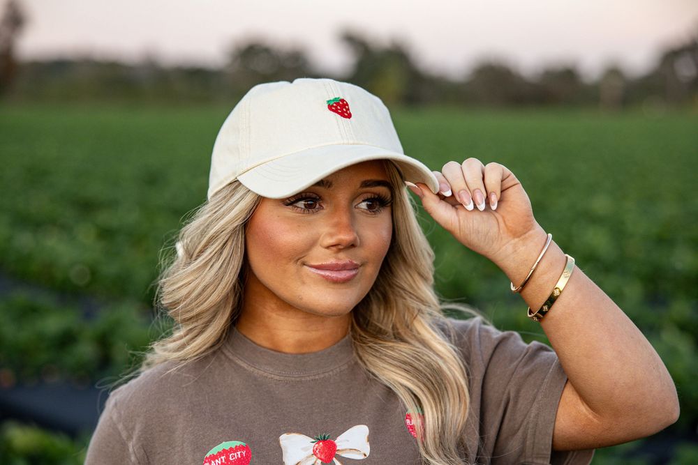Woman wearing a cream baseball cap with strawberry embroidery and a graphic tee, posing in a green field.