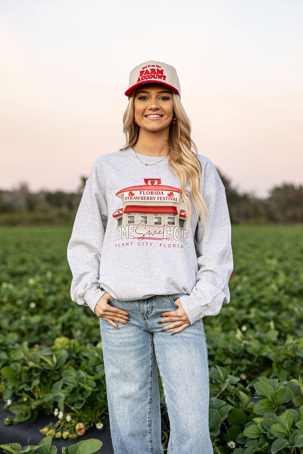 Woman in gray Florida Strawberry Festival sweatshirt and red Farm Account cap standing in a green field.