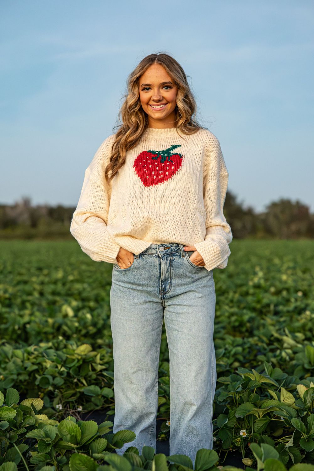 Woman in cream strawberry sweater and jeans standing in a green field under a blue sky.