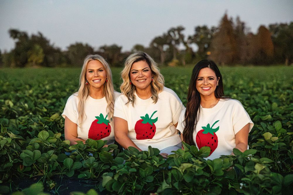 Three women wearing strawberry graphic tees smiling in a lush green strawberry field at golden hour.