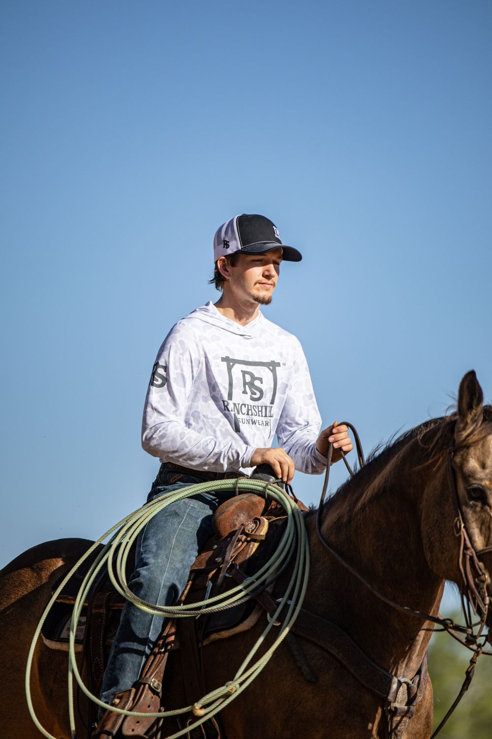 Young cowboy on horseback holding a lasso rope, wearing a white hoodie and cap against a clear blue sky.