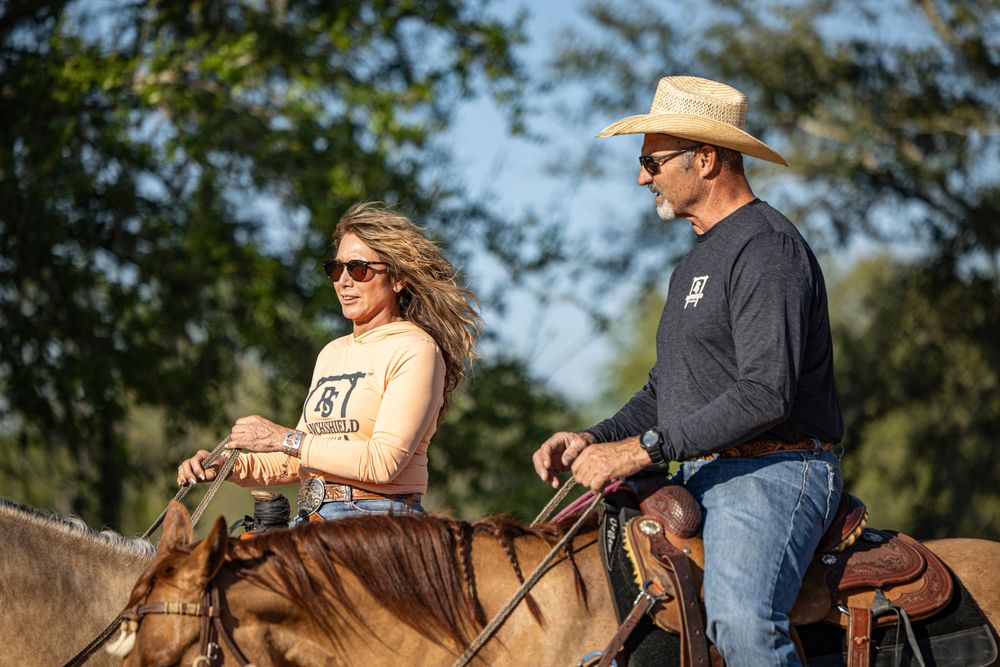 Two people riding horses outdoors wearing cowboy hats and sunglasses on a sunny day with green trees in background.