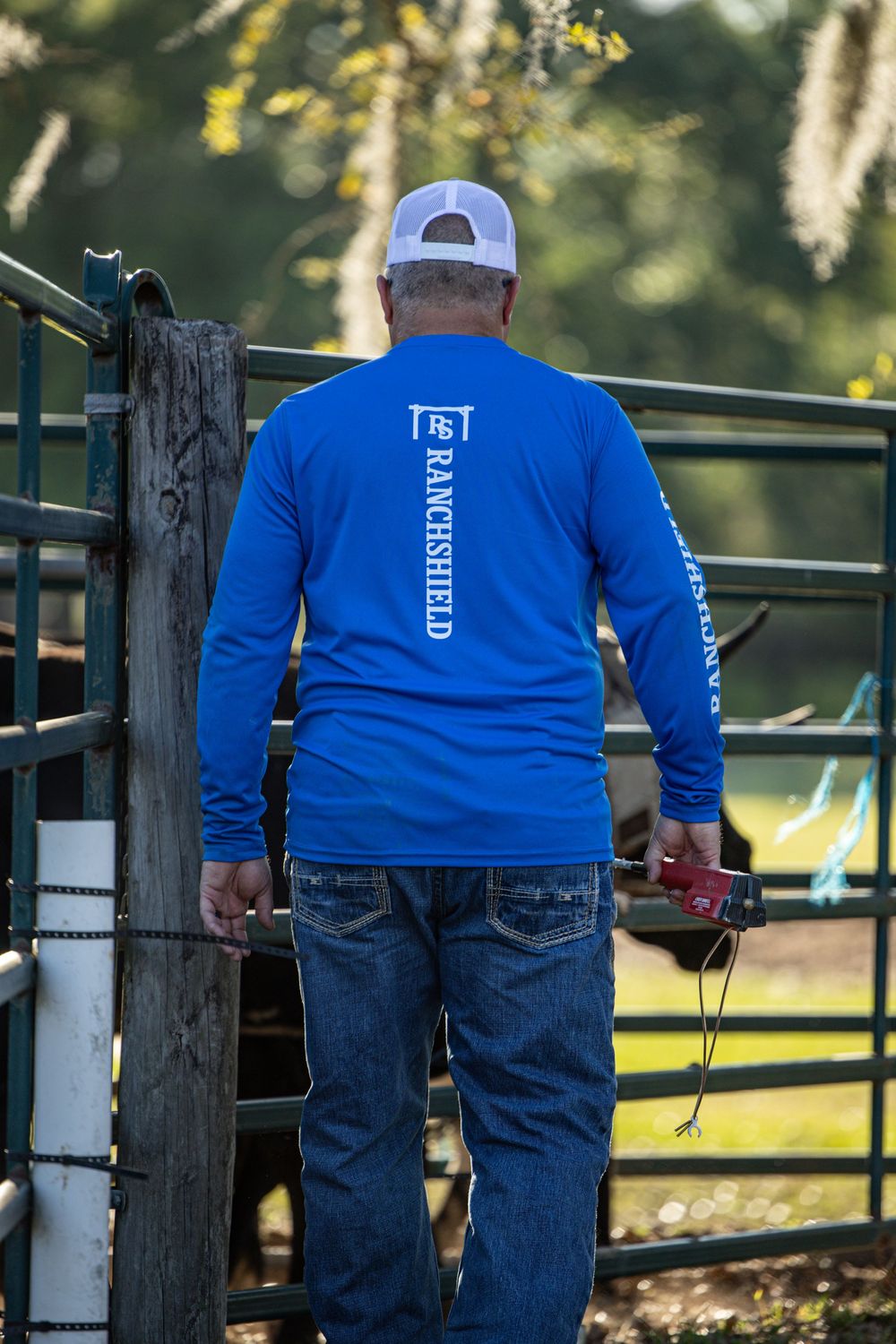 Man in blue RanchShield long sleeve shirt and white cap standing at metal cattle pen gate on a ranch.