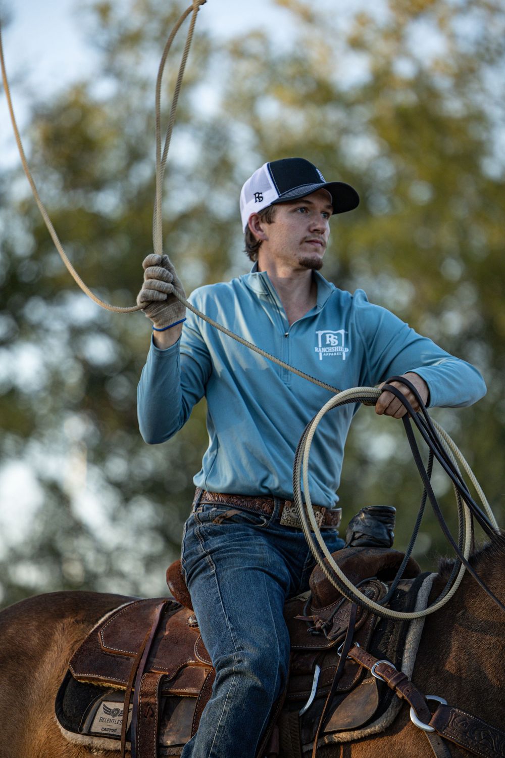 Cowboy on horseback holding a lasso rope, wearing a blue shirt and trucker hat, practicing roping outdoors.